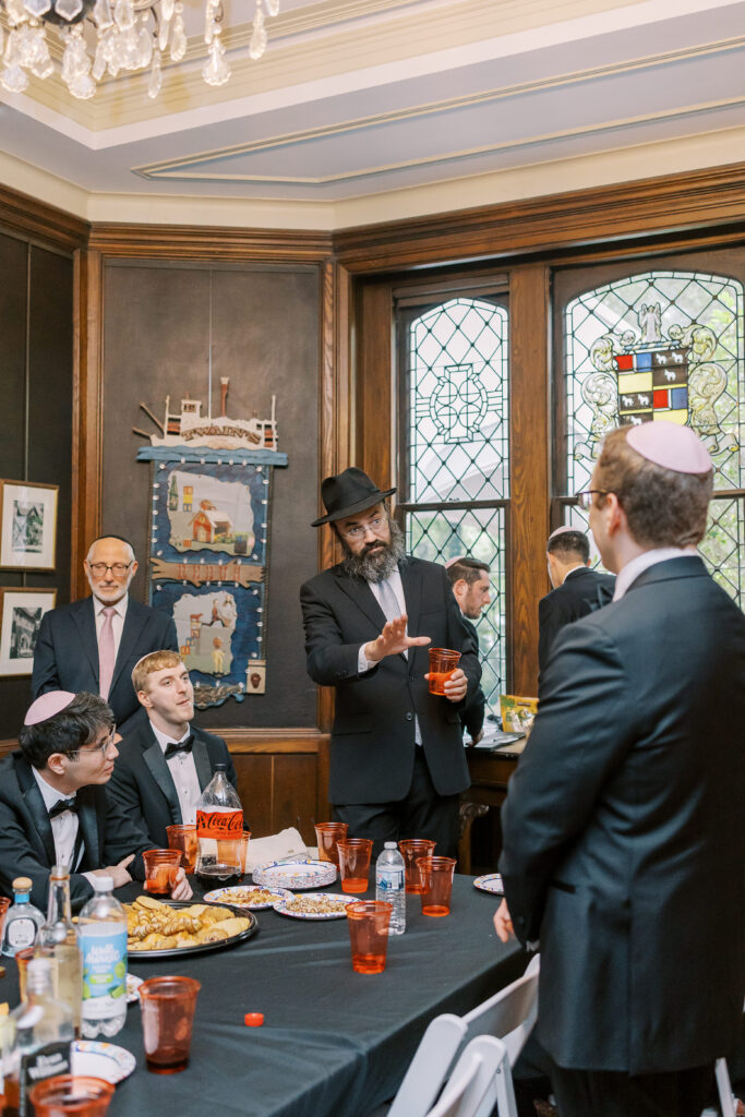 A group of men, some wearing yarmulkes, gather around a table with food and drinks from kosher wedding catering Atlanta in a wood-paneled room. One man stands gesturing as others listen. Stained glass windows and wall art decorate the background.