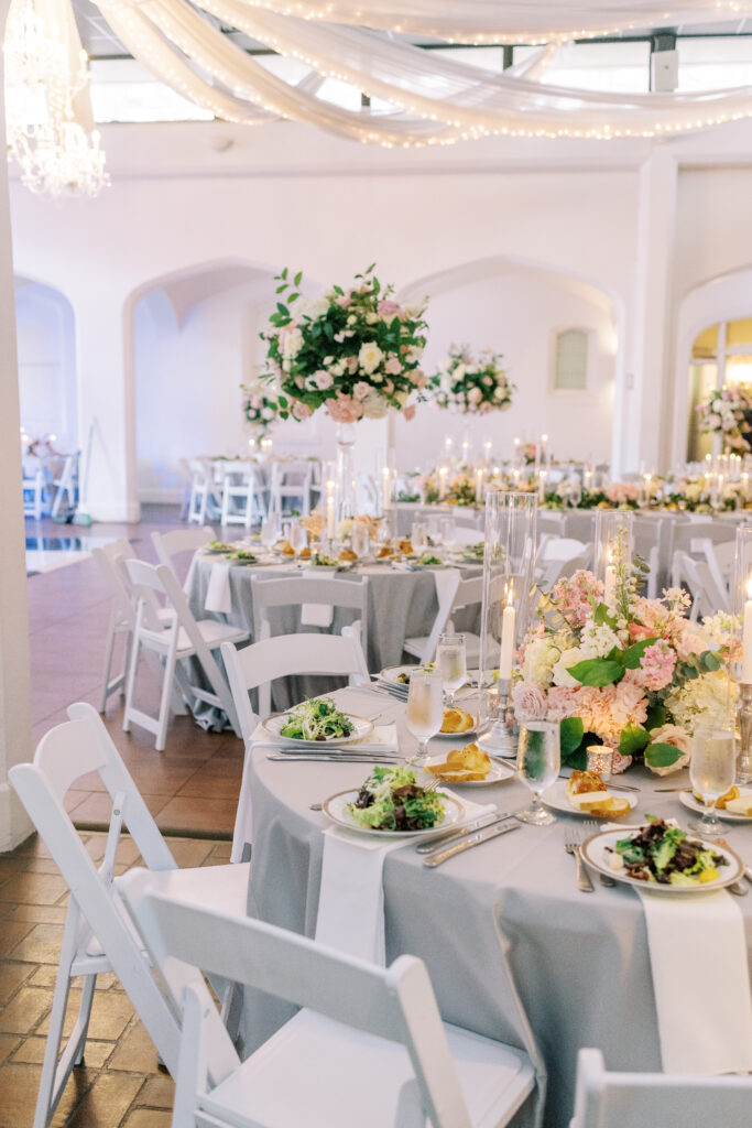 A beautifully decorated wedding reception with white chairs, round tables with gray tablecloths, elegant floral centerpieces, plates of salad from top-notch kosher wedding catering Atlanta, and soft candlelight under draped fabric and string lights.