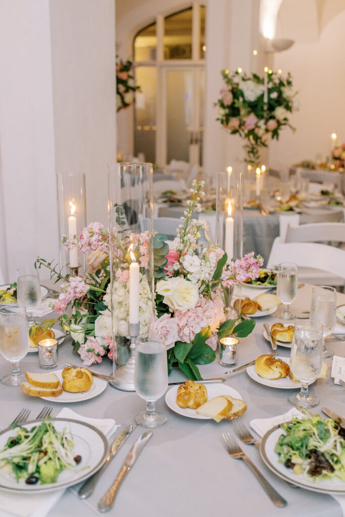 A round table set for a formal event with salads, rolls, water glasses, candles, and a large floral centerpiece of white and pink flowers—perfectly complemented by expert kosher wedding catering Atlanta. White chairs and more decorated tables are visible in the background.