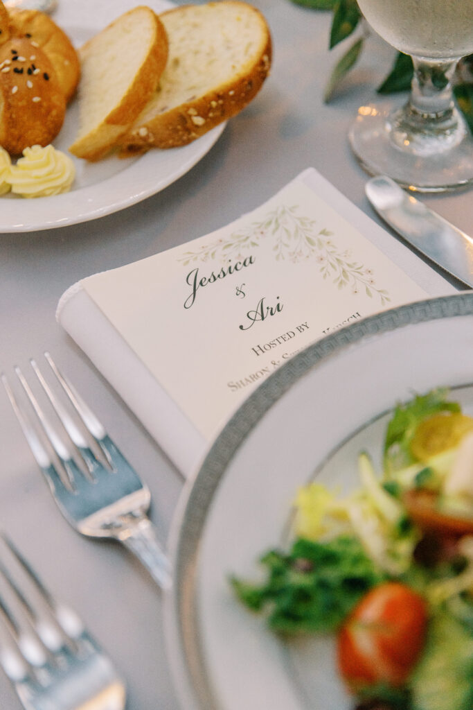 A close-up of a wedding menu with Jessica & Ari on the cover, next to a plate of salad, bread with butter, silverware, and a glass of water on an elegantly set table—showcasing the attention to detail provided by kosher wedding catering Atlanta.