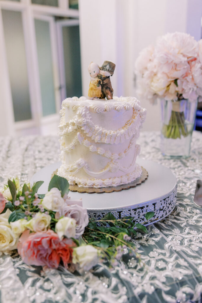 A white frosted cake with piped icing and a porcelain couple topper sits on a silver stand next to a bouquet of flowers; pink and white blooms, thoughtfully arranged by expert kosher wedding catering Atlanta professionals, are in a vase in the background.