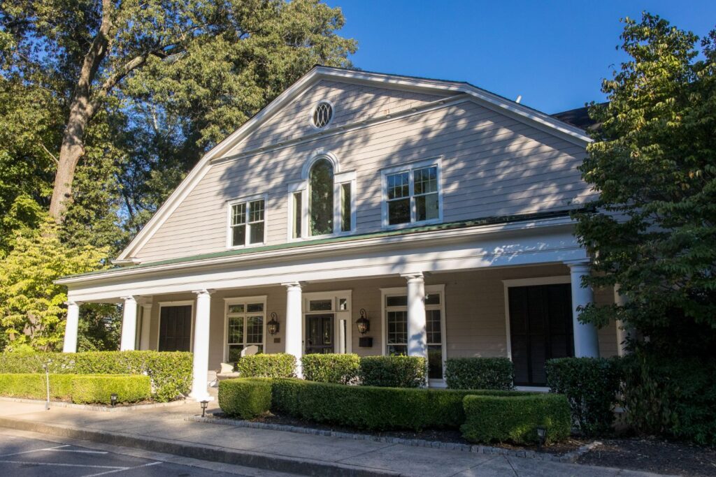 Light gray two-story house with white columns, porch, and manicured bushes in front. Large trees provide shade, and sunlight highlights part of the house against a clear blue sky.