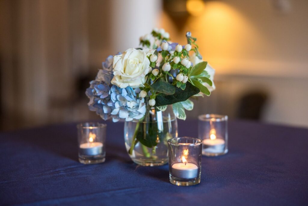 A small glass vase with white roses and blue hydrangeas sits on a dark blue tablecloth, surrounded by three lit tealight candles in glass holders.