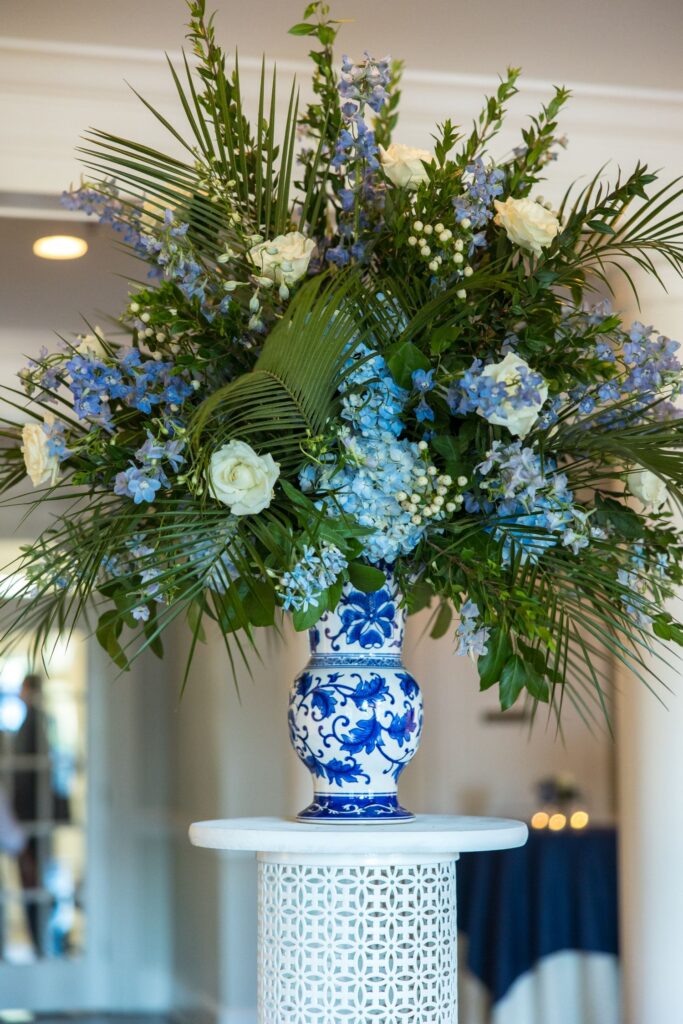 A blue and white porcelain vase holds a large floral arrangement of white roses, blue flowers, and assorted green foliage, placed on a white pedestal indoors.