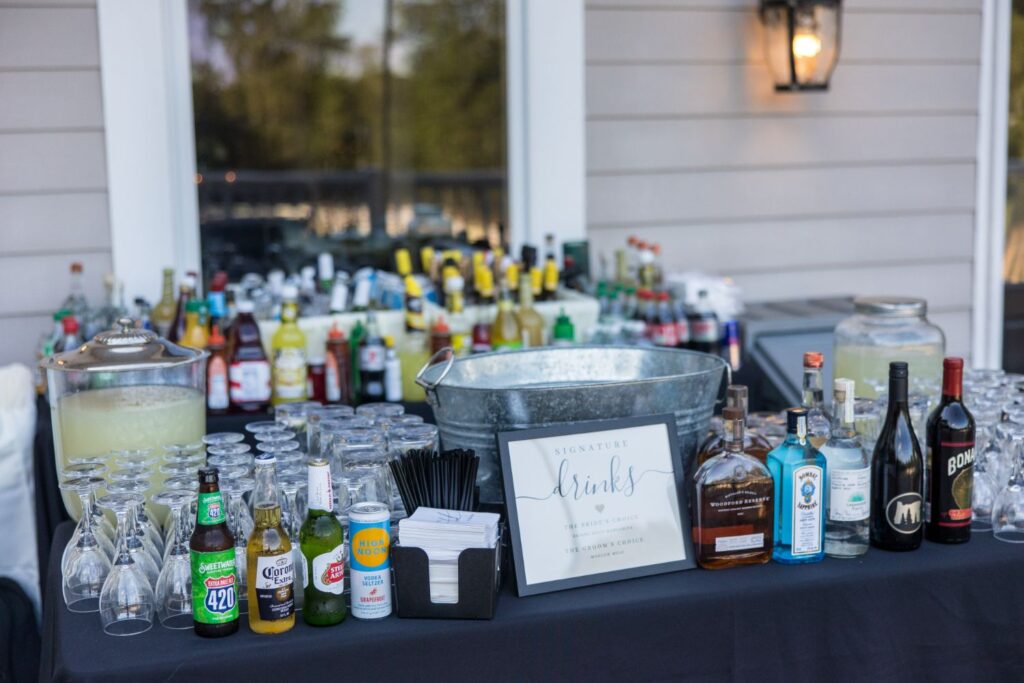 Outdoor bar setup with assorted liquor bottles, mixers, glassware, a metal tub, drink dispensers with lemonade, canned drinks, cocktail napkins, black straws, and a framed Signature Drinks sign on a black tablecloth.