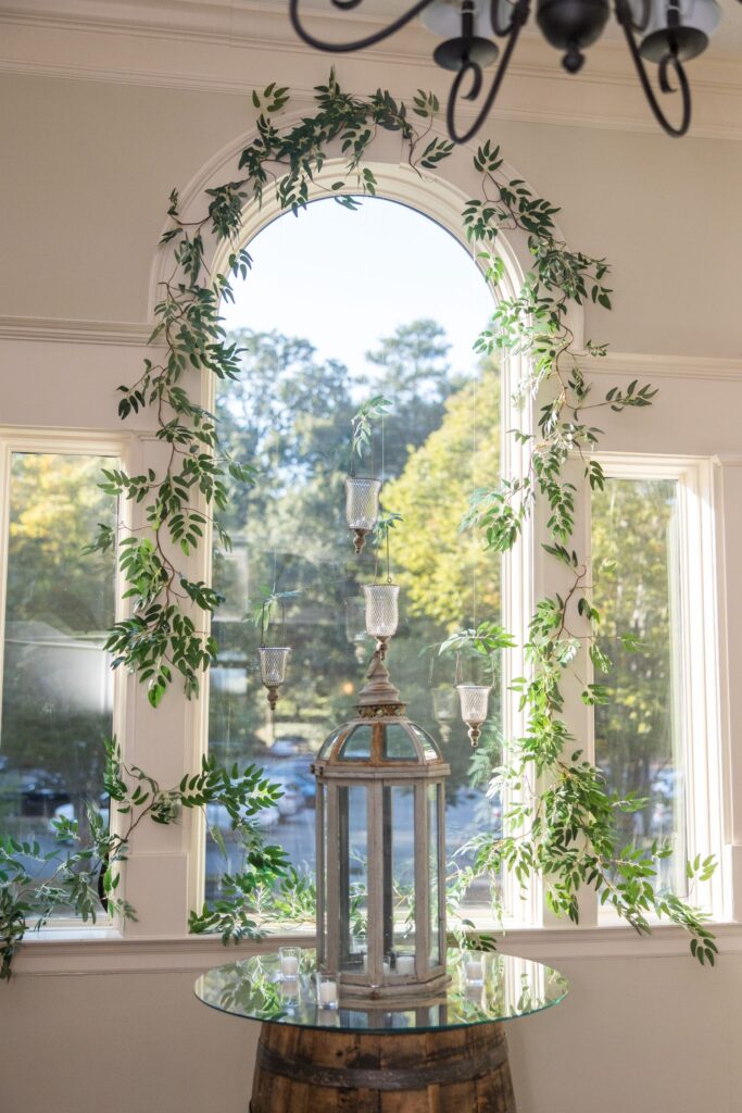 Large arched window framed by greenery, with a decorative lantern and hanging glass candle holders displayed on a glass-topped barrel inside a bright room. Trees and sky are visible through the window.