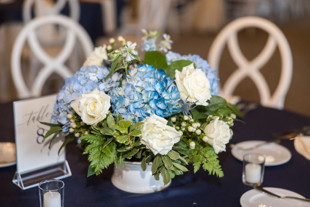 A floral centerpiece with blue hydrangeas, white roses, and greenery sits on a round table with white chairs, a table number sign, plates, cutlery, and small candles.