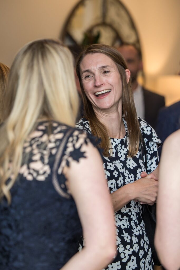 A woman in a black and white floral dress smiles and talks with another woman wearing a dark lace dress at a social gathering.