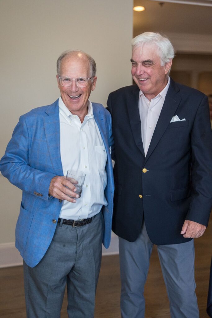 Two older men in blazers stand smiling together indoors. One man holds a drink and wears a blue jacket, while the other wears a dark blazer with a white shirt and pocket square.