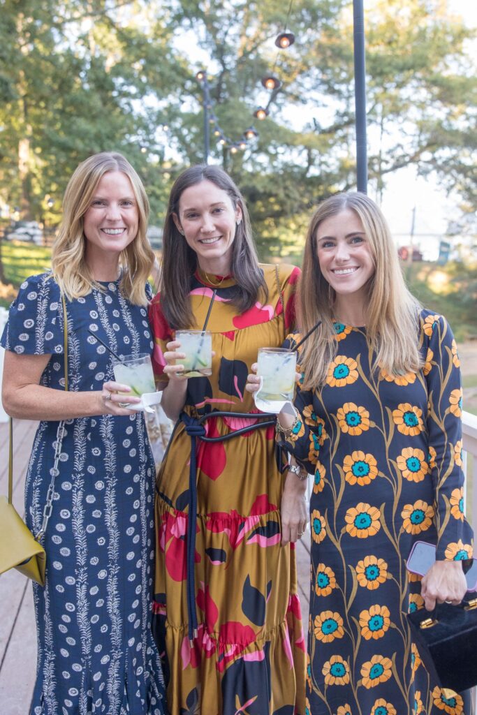 Three women stand outdoors, smiling and holding drinks. They wear long, colorful dresses with floral and patterned designs. Trees and string lights are visible in the background.