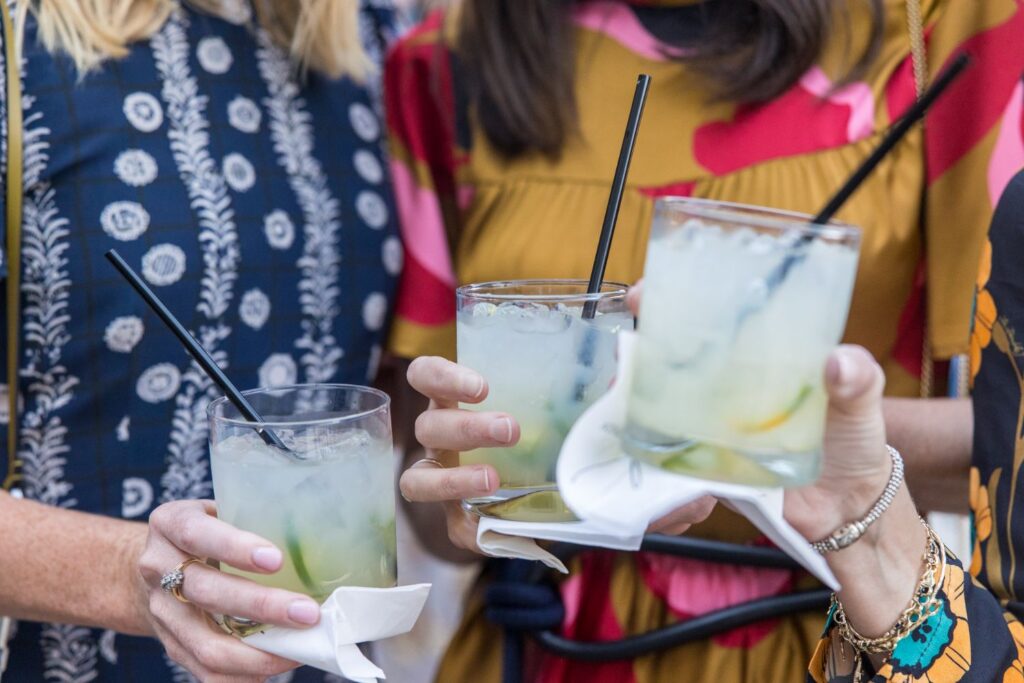 Three women in colorful patterned dresses hold cocktails with lime and black straws, each resting on a white napkin. The image is cropped to show only their hands and torsos.