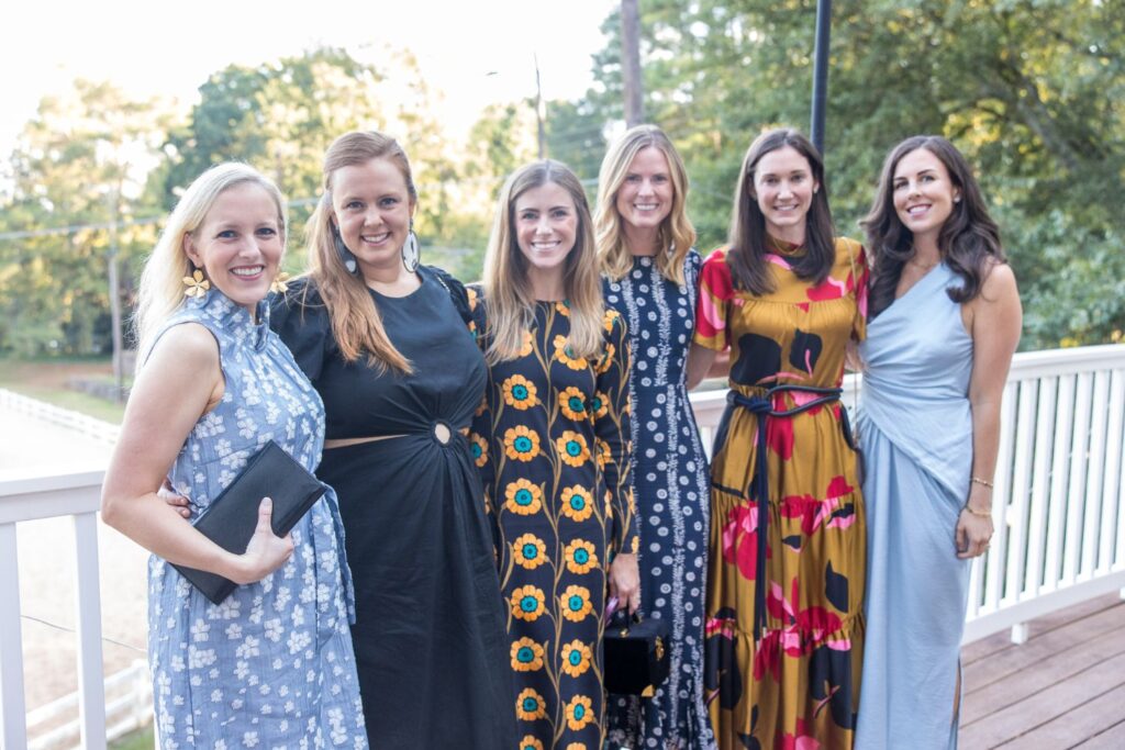 Six women stand together, smiling on a wooden deck with trees in the background. They are dressed in colorful, patterned dresses, and appear to be at an outdoor event or gathering.