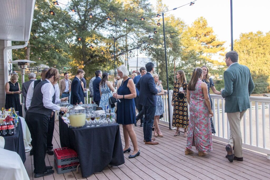 People in semi-formal attire gather on a wooden deck for an outdoor event, chatting and enjoying drinks by a beverage table, with string lights overhead and trees in the background.