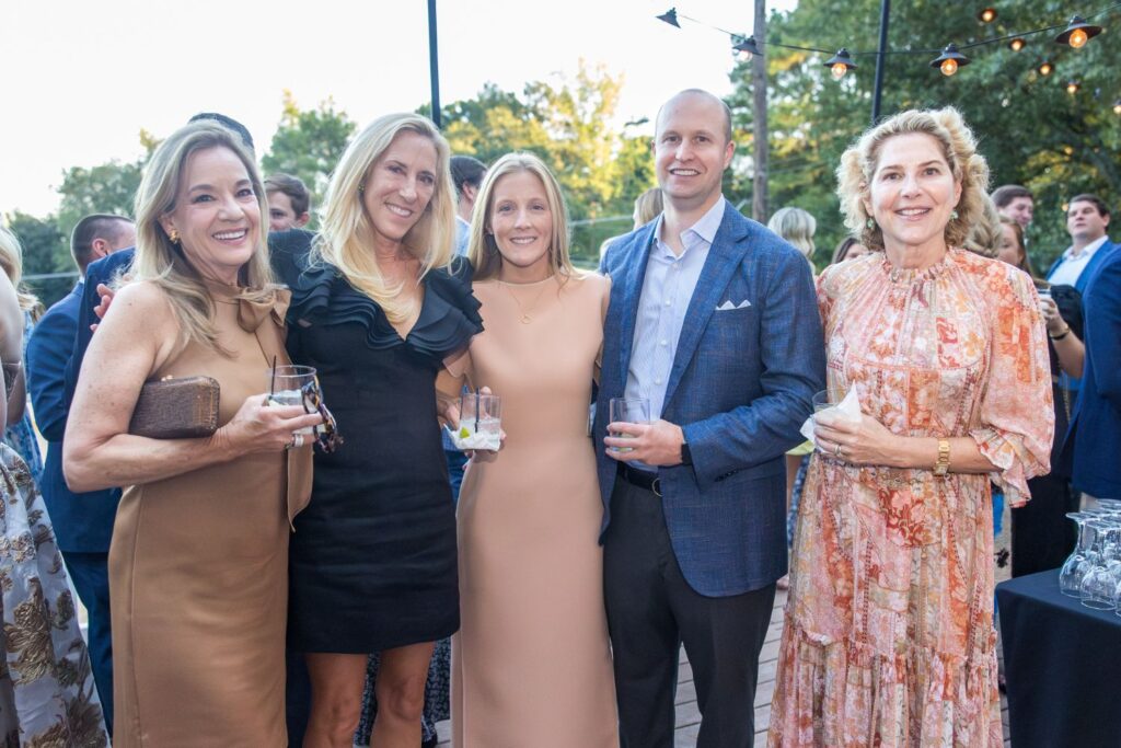 Five people stand close together at an outdoor event, smiling and holding drinks. Three women wear dresses, one man is in a suit, and another woman wears a floral dress. String lights and trees are visible in the background.