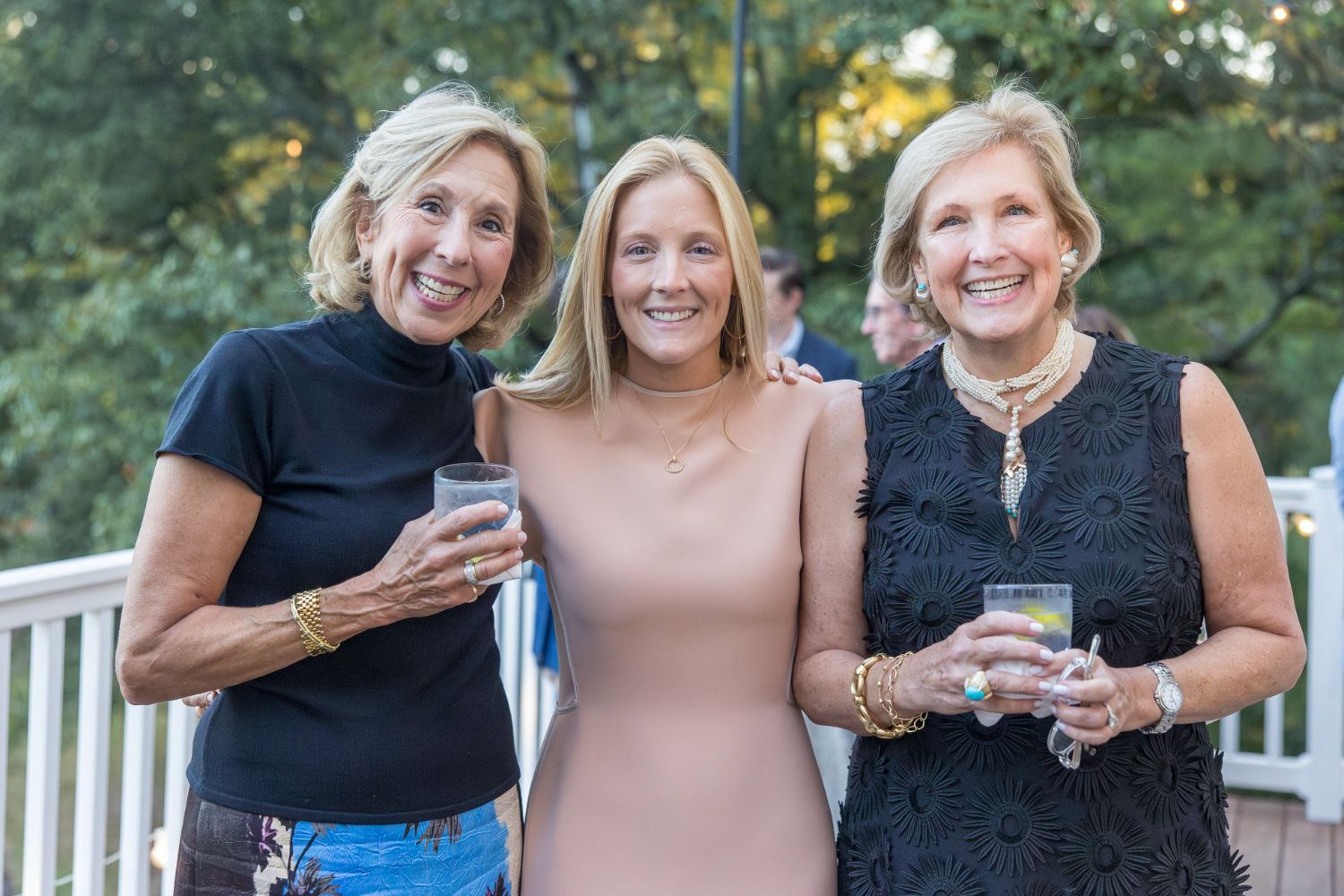 Three women smile and pose together outdoors on a deck, holding drinks. Trees and other people are visible in the background. The women are dressed in a mix of formal and casual attire.