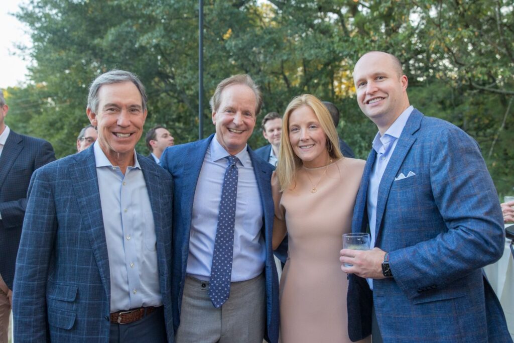 Four people dressed in formal attire smile and pose together outdoors at a social event, with greenery and other guests visible in the background.