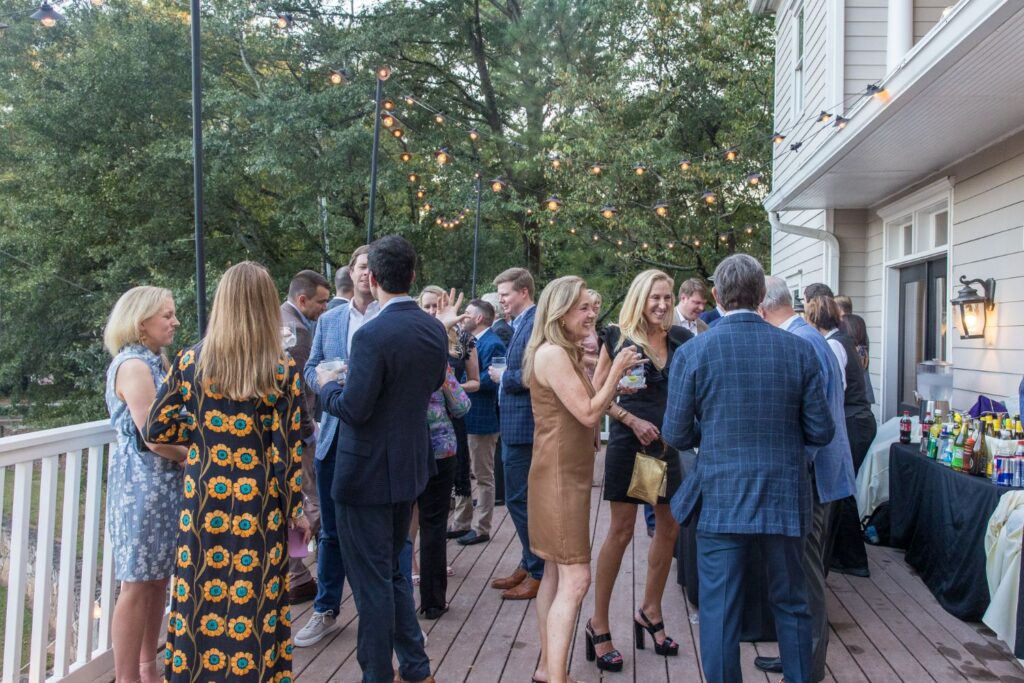 A group of people in semi-formal attire mingle and chat at an outdoor event on a wooden deck with string lights and trees in the background. A drink table is set up along the house wall.