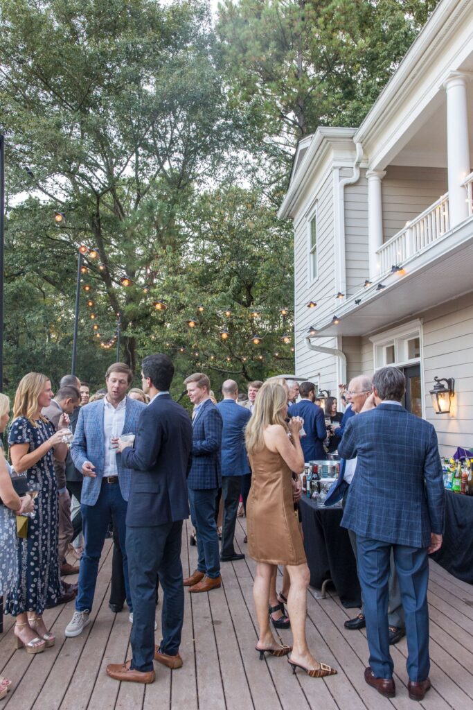 People in semi-formal attire socialize on a wooden deck at an outdoor gathering beside a light-colored house, with trees and string lights in the background.
