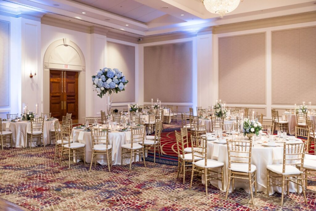 A kosher wedding reception in Atlanta held in a banquet hall with round tables covered in white linens, gold-framed chairs, and centerpieces featuring blue and white hydrangeas.