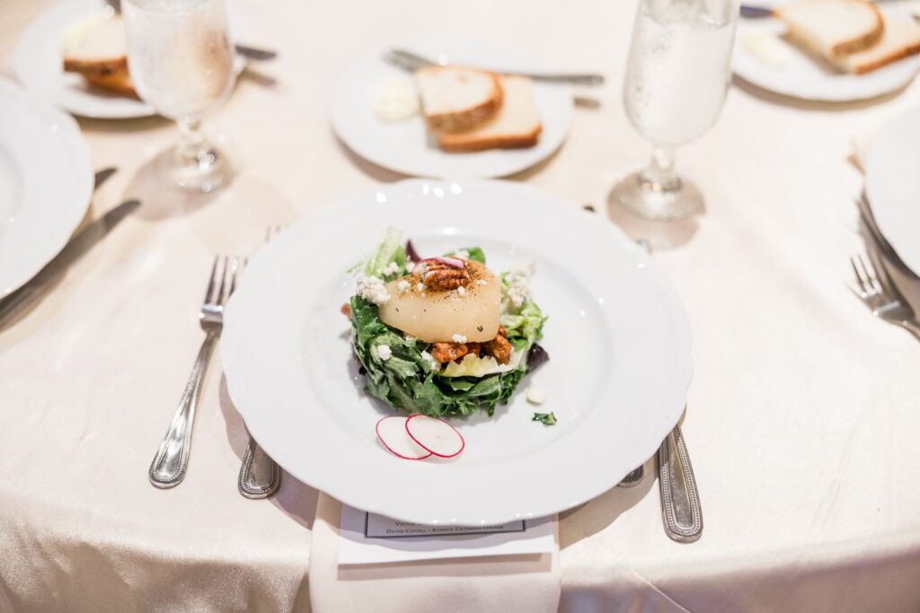 A salad topped with a poached pear, nuts, and cheese on a white plate, served at a kosher wedding reception in Atlanta with slices of bread and water glasses set on a table.