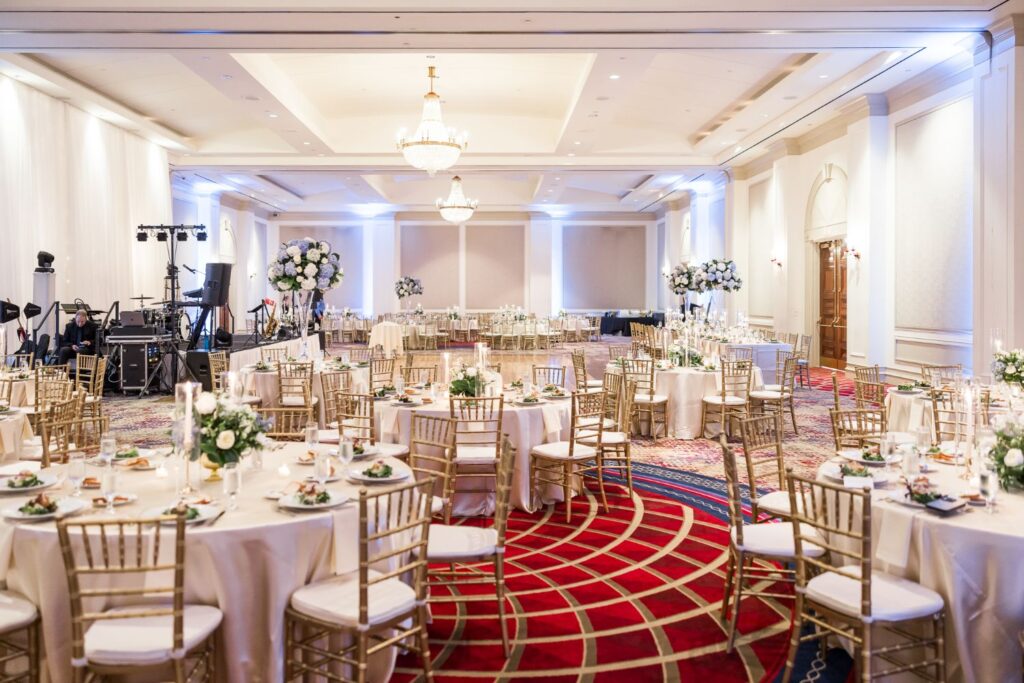 A large banquet hall in Atlanta set for a kosher wedding reception with round tables, gold chairs, and floral centerpieces atop a patterned red carpet.