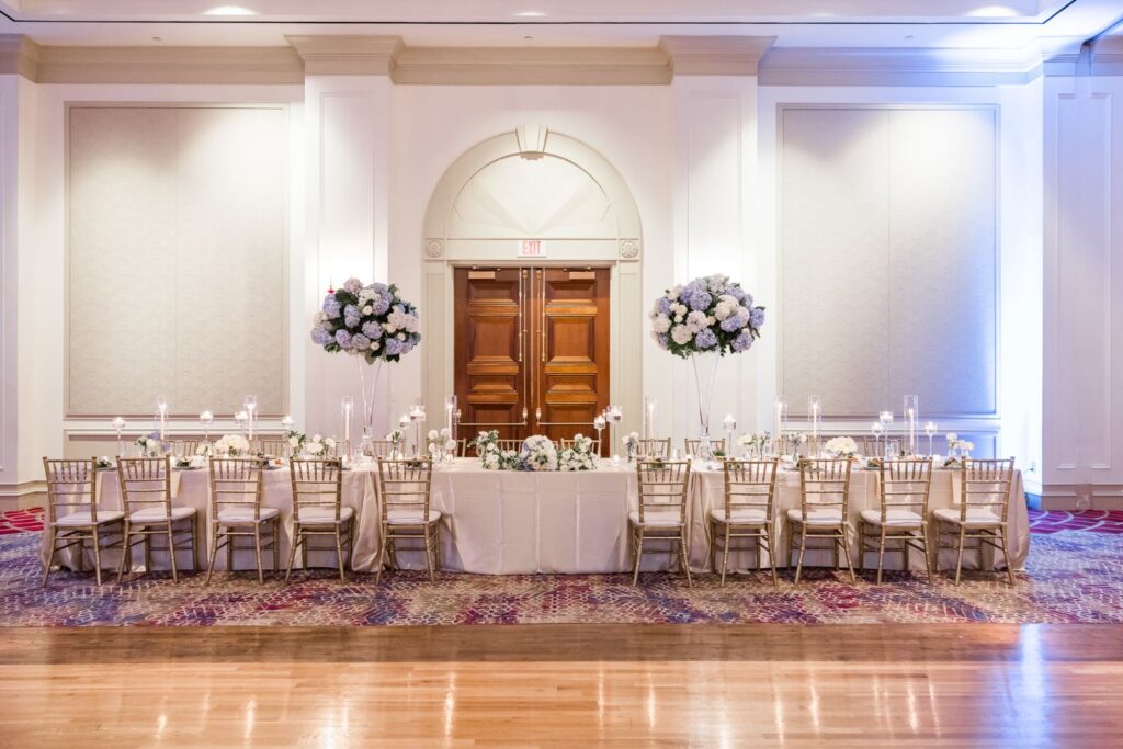 Long rectangular wedding head table with cream linens, gold chairs, and tall floral centerpieces in a formal Atlanta ballroom set for a kosher wedding reception.