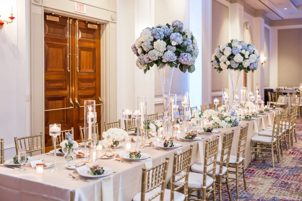 A long banquet table set for a formal Atlanta kosher wedding reception, decorated with tall glass centerpieces featuring blue and white hydrangeas and surrounded by gold chairs.