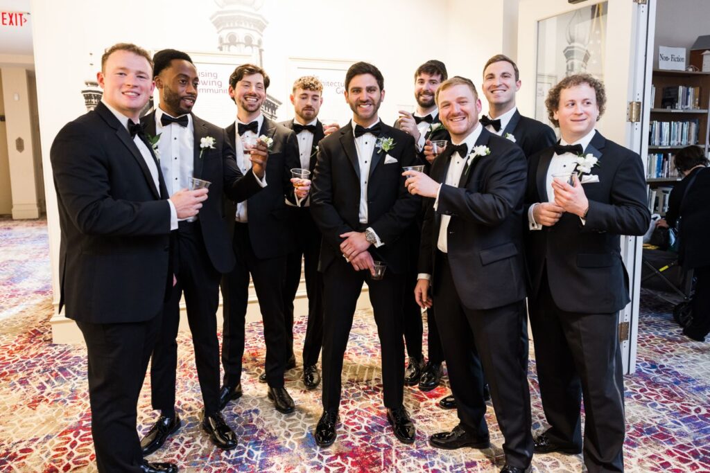 A group of nine men in black tuxedos hold small clear cups and smile during a kosher wedding reception in Atlanta, standing in a room with patterned carpet.