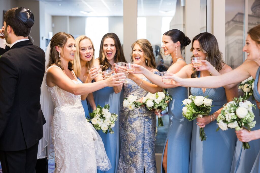 A bride in a white wedding gown and five bridesmaids in blue dresses toast with clear plastic cups at an Atlanta kosher wedding reception.
