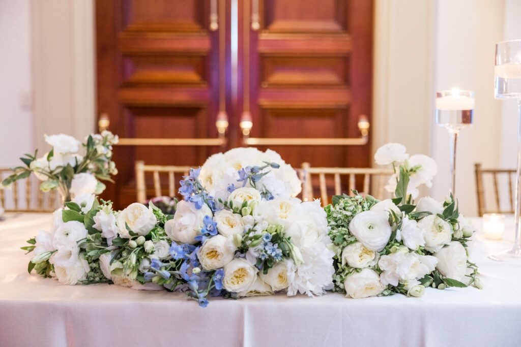 A long floral centerpiece of white roses, peonies, and blue delphiniums arranged on a table in front of double wooden doors at a kosher wedding reception in Atlanta.