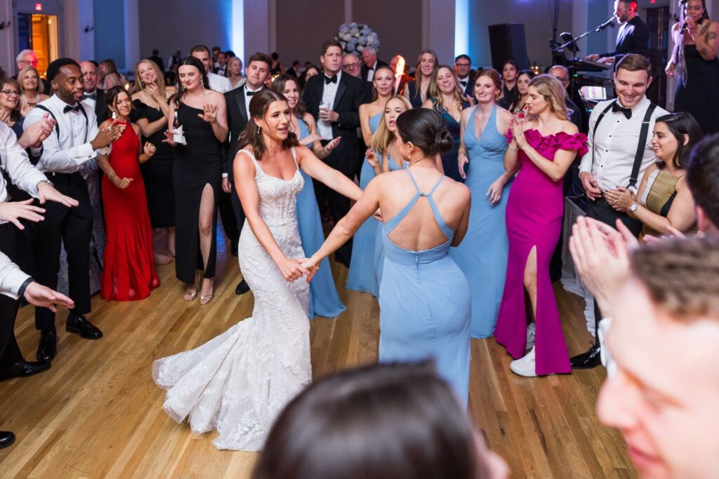 A bride in a white wedding gown dances with a bridesmaid in a blue dress, surrounded by wedding guests on a wooden dance floor at a kosher wedding reception in Atlanta.