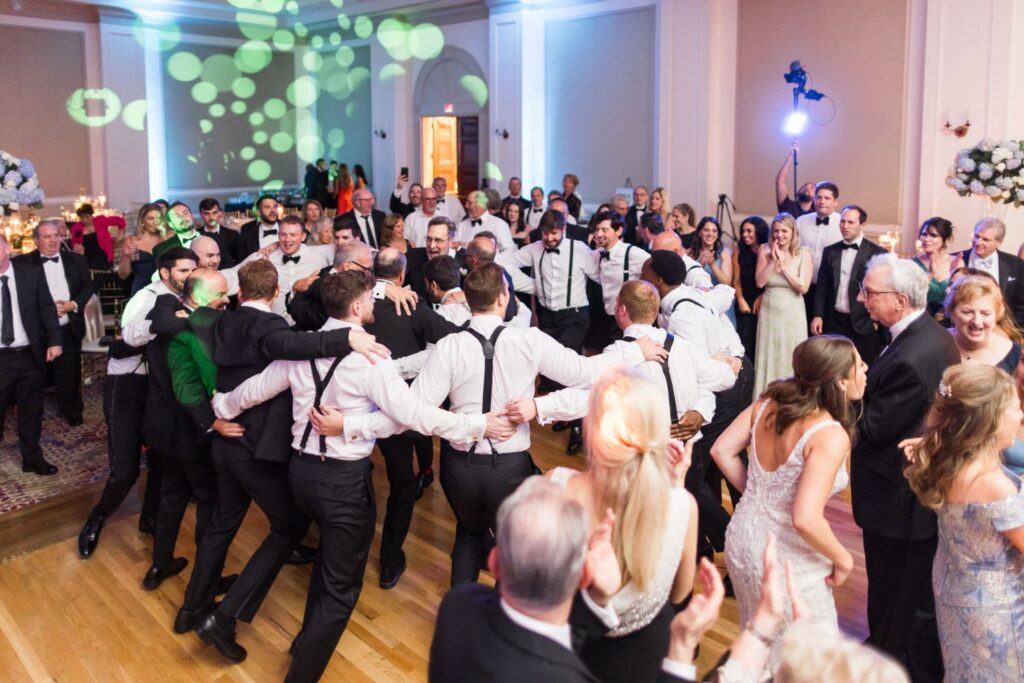 Wedding guests in formal attire dancing in a large ballroom at a kosher wedding reception in Atlanta, with a group of men in white shirts and suspenders linked together in the center.
