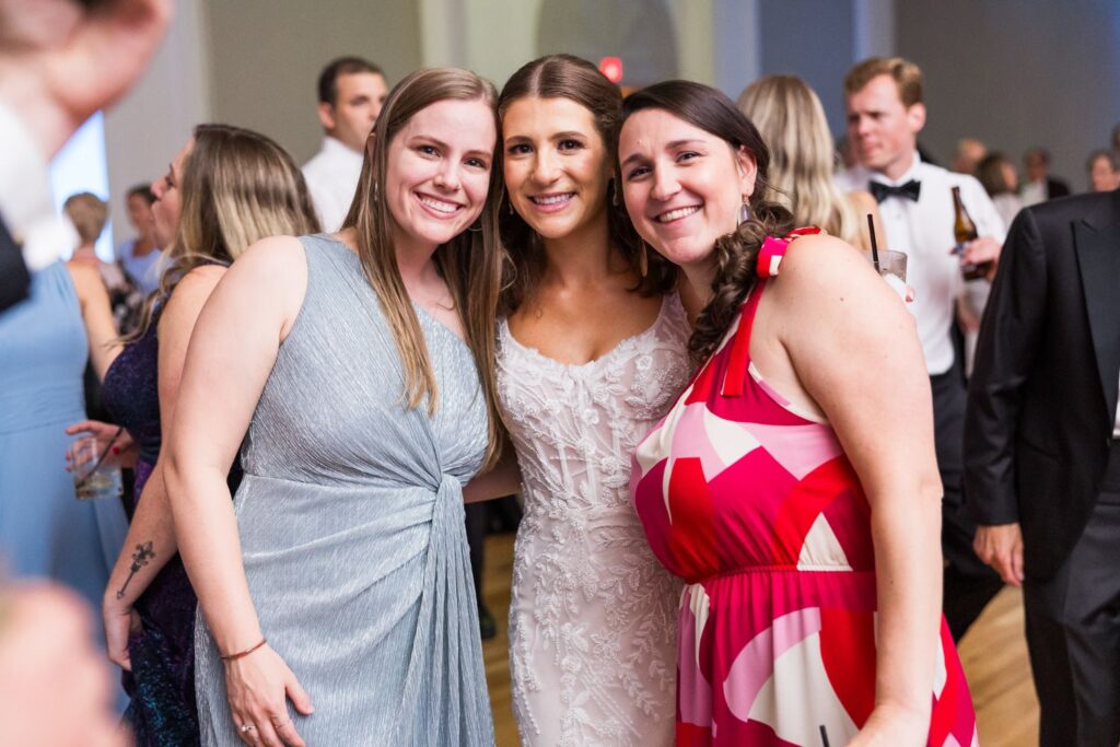 Three women smile and pose for a photo at a kosher wedding reception in Atlanta, with the bride in the center wearing an embellished white gown.