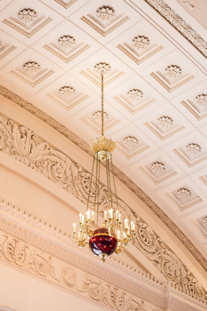 Ornate brass chandelier with a large red glass orb, suspended from a coffered ceiling with decorative molding above a kosher wedding reception in Atlanta.