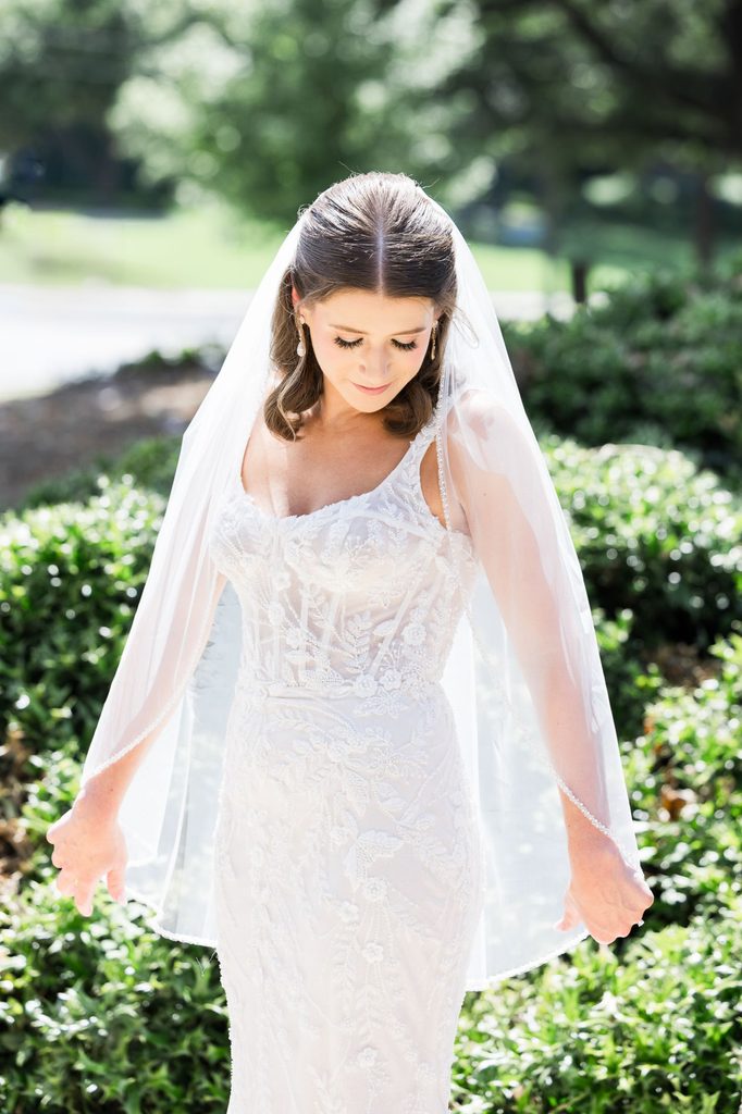 A bride in a beaded, corset-style wedding gown and veil looks down at her kosher wedding reception in Atlanta, holding the edges of her veil.