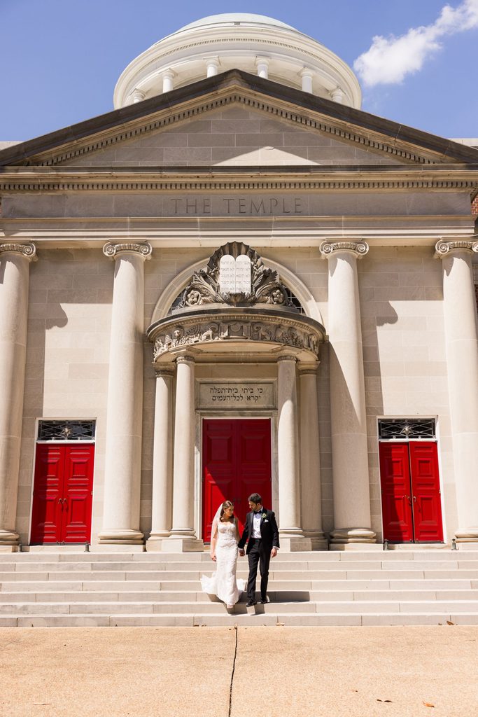 A bride and groom hold hands while walking down the steps in front of The Temple, the site of a kosher wedding reception in Atlanta with bright red doors.
