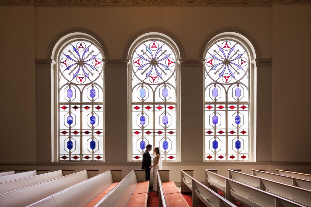 A bride and groom stand facing each other in the aisle of a church during a kosher wedding reception in Atlanta, framed by three large, arched stained-glass windows.