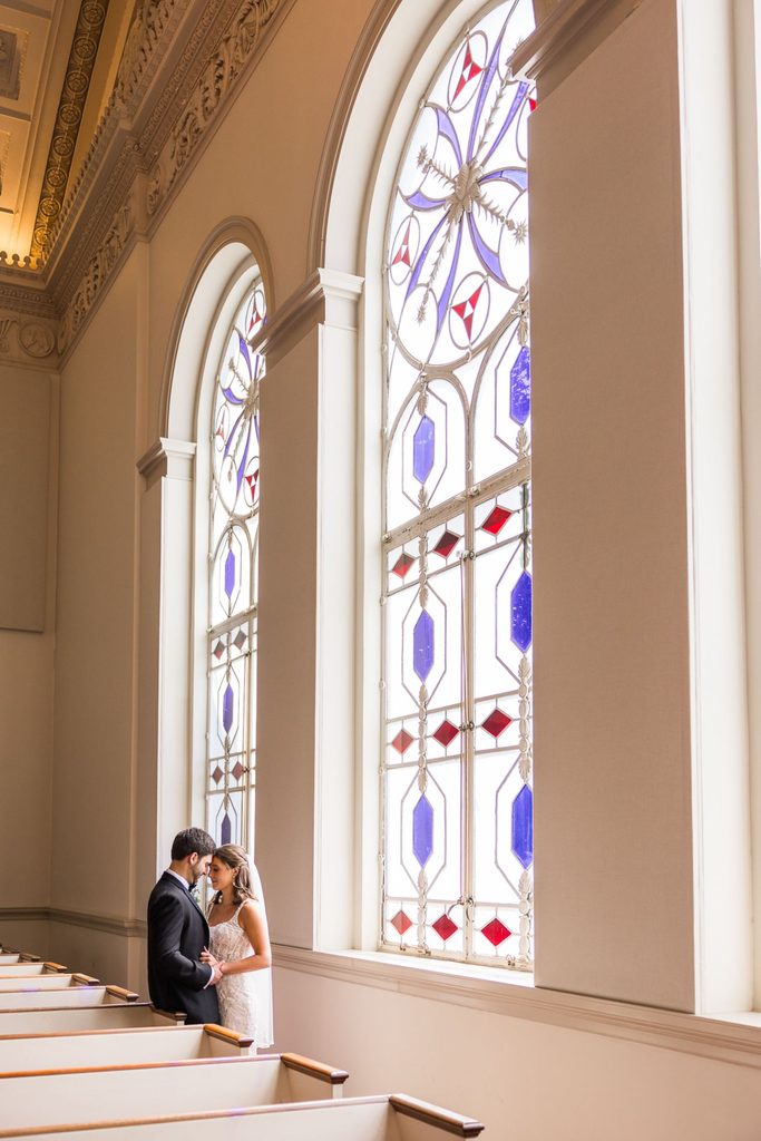 A bride and groom stand forehead-to-forehead in a church aisle next to tall, ornate stained-glass windows at their Atlanta wedding reception.