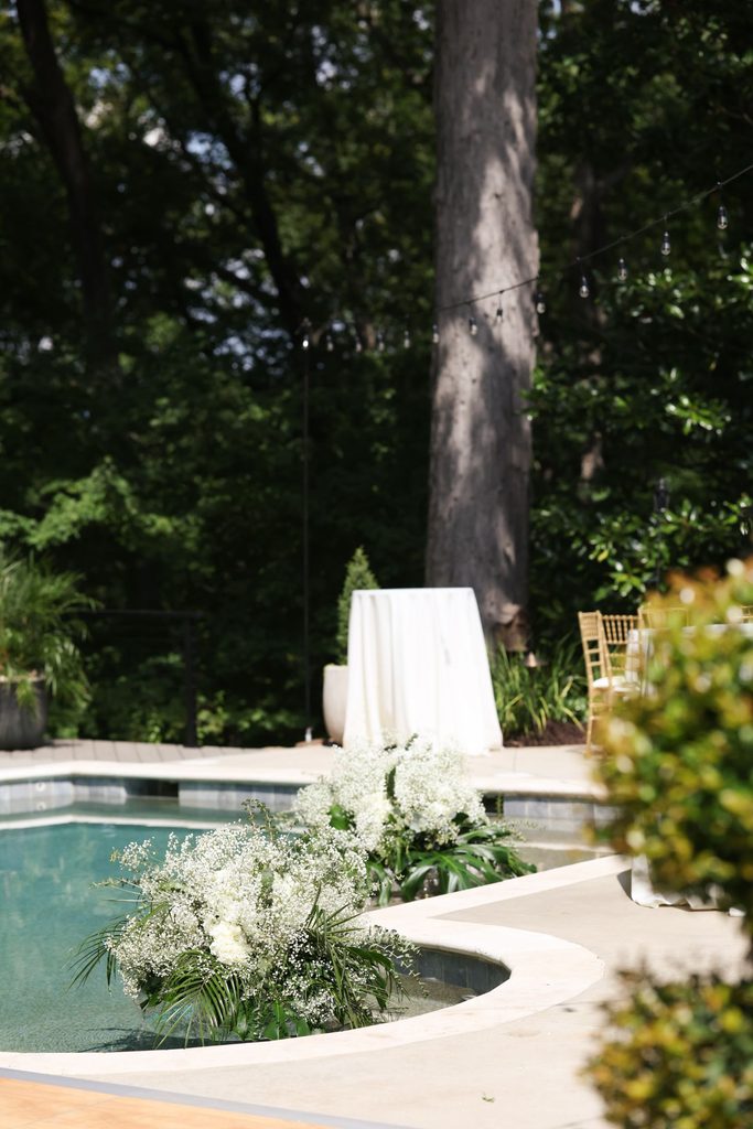 Large floral arrangements of white babys breath sit on the edge of a swimming pool, with a draped cocktail table and gold chairs in the background.