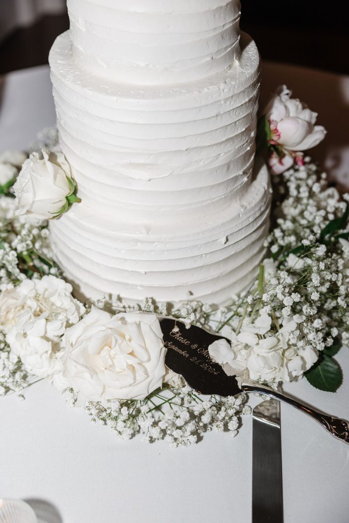 A two-tiered white frosted cake surrounded by white roses and babys breath, with a personalized cake server resting at the base.