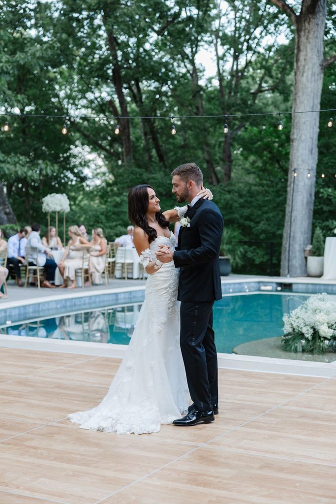 A bride and groom share their first dance on a wooden dance floor in front of a backyard swimming pool.