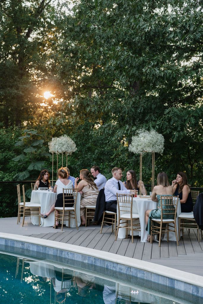 Guests seated at round tables on a poolside deck during an outdoor evening event.