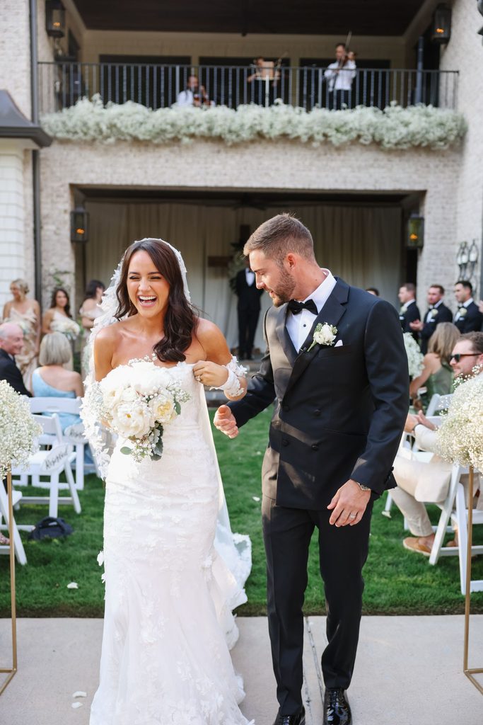 A bride in a white wedding gown and a groom in a black tuxedo walking down the aisle together after their ceremony, both laughing.