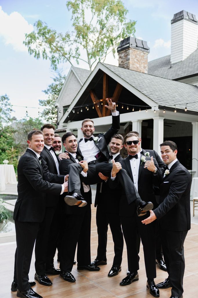 A group of eight groomsmen in black tuxedos lifting the groom above their heads outdoors.