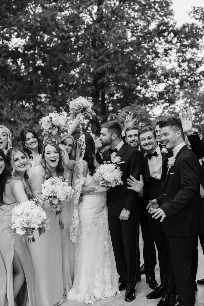 A bride and groom kissing, surrounded by their wedding party outdoors.