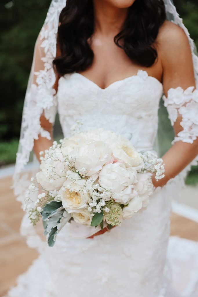 A bride in a strapless lace wedding dress and veil holds a bouquet of white peonies and babys breath.