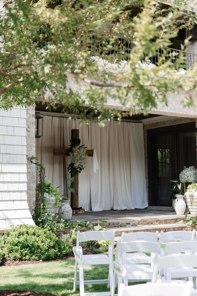 Wooden cross decorated with white flowers on a patio covered by cream curtains, viewed from behind rows of white folding chairs.