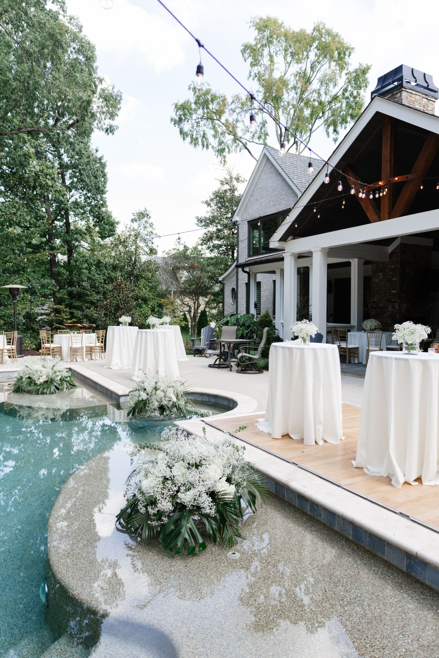 Outdoor patio with high-top tables covered in white tablecloths set up around a swimming pool decorated with floral arrangements.