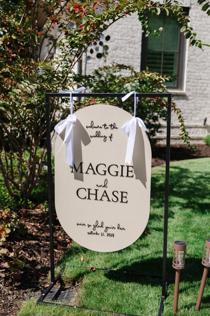 Oval wedding welcome sign on a black metal stand reading: Welcome to the wedding of Maggie and Chase, were so glad youre here, September 21, 2023.