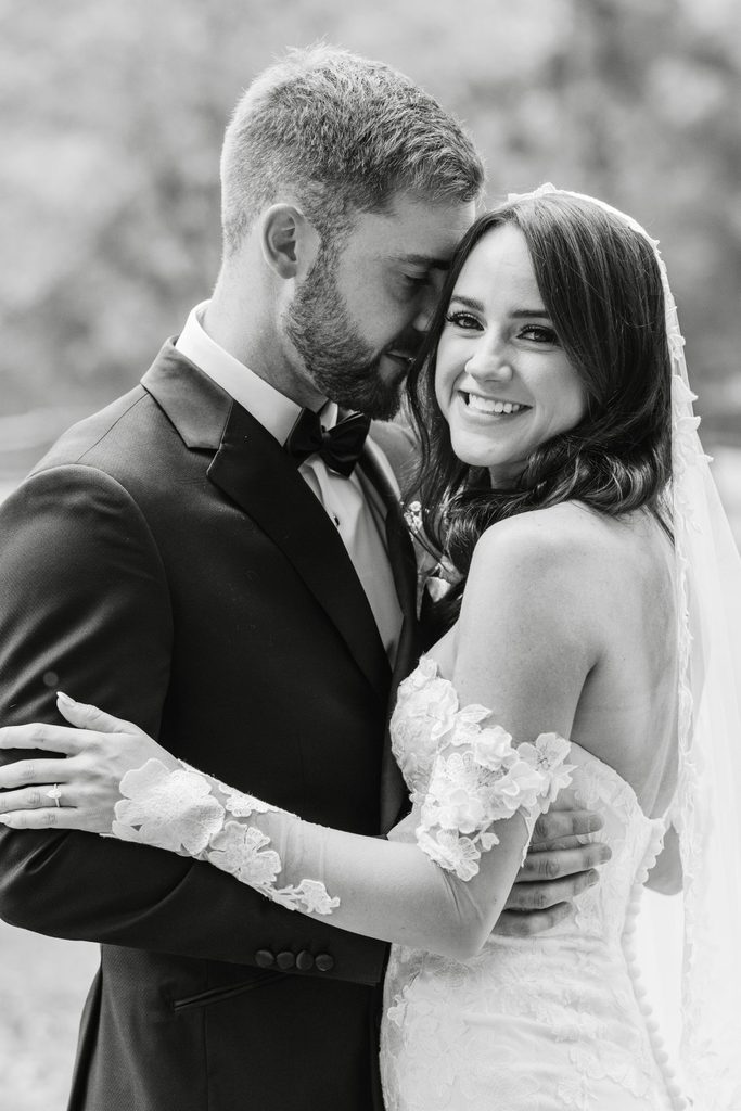 A bride in an off-the-shoulder lace gown and a groom in a tuxedo embrace closely, smiling at the camera.